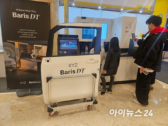 시연에 나선 직원이 25일 제주 신화월드에서 열린 국제 e-모빌리티 엑스포 부스에서 의자에 앉아 창문 너머로 음료를 받고 있다. [사진=설재윤 기자]